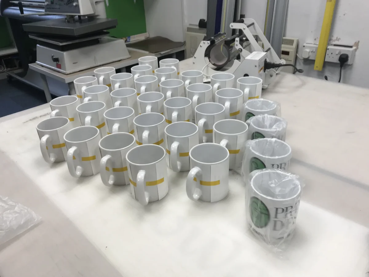 Rows of ceramic mugs beside heat-press equipment