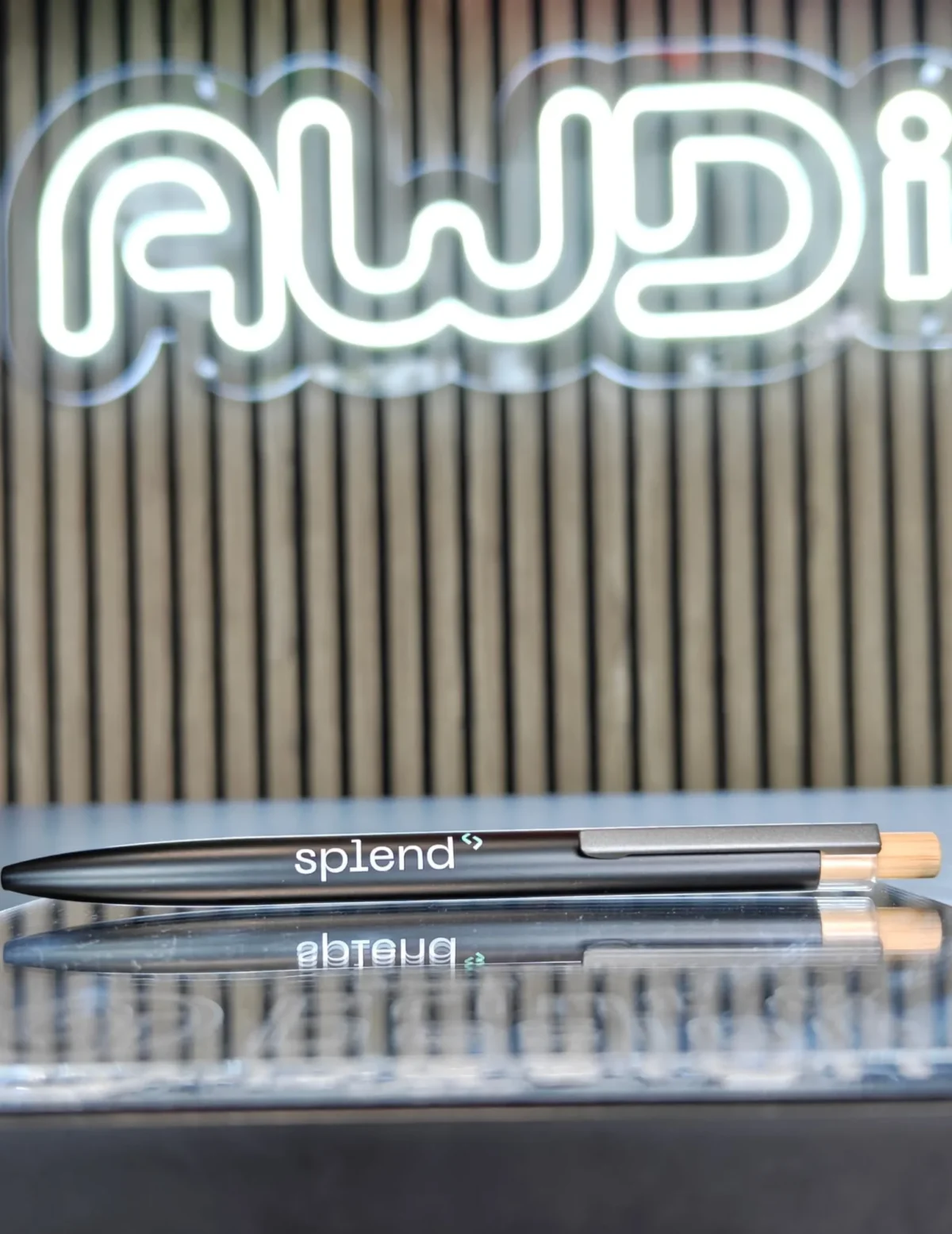 Black Splend branded pen on reflective surface beneath neon sign